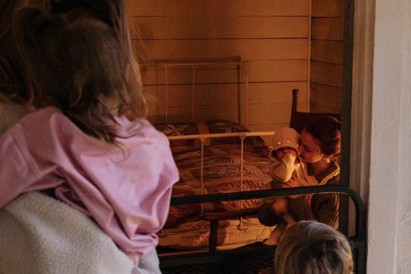 A mom and two young girls look inside the box and strip house, where a young mom and baby are rocking in a rocking chair.
