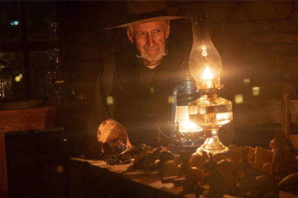 A bearded cowboy sits with an oil lamp. Several hand-carved toys are on the table in front.