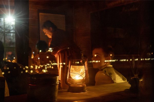 A lamp luminates the inside of the one-room schoolhouse. Children are sitting at their desk learning lessons.
