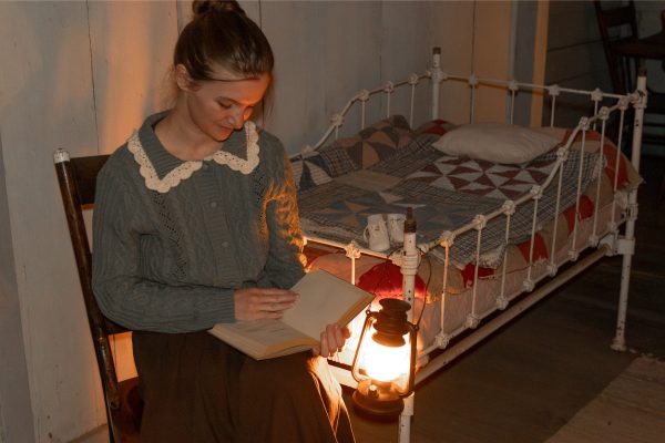 A young woman reads a book by lamp. A small child's bed is in the background with a quilt on top.