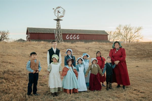 A group photo of 10 young school children with their teacher. All are volunteers dressed in period clothing of the early 1900s. In the background is a windmill and the side of the Four Sixes horse barn.