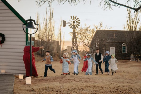 Young school children running to their teacher outside the one-room school house. The teacher is ringing a bell and there is a windmill in the background.