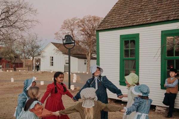 The young school girls playing ring around the rosie outside of the one-room school house.
