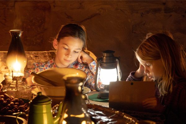 Two young girls dressed in 1800s period clothing reading books by lamp light.