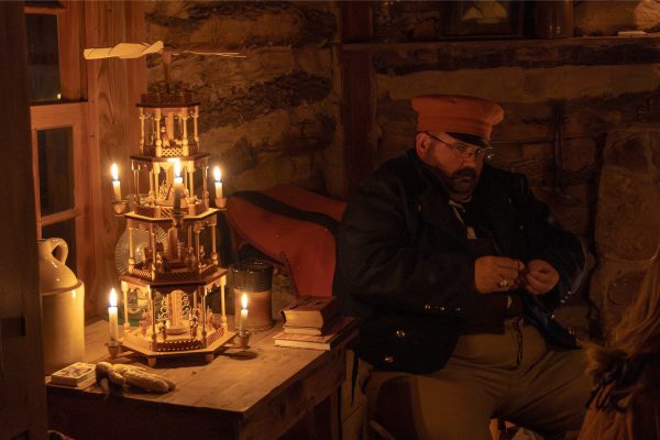 A man dressed in German period clothing from the 1800s sits next to a German Christmas carousel lit with candles.