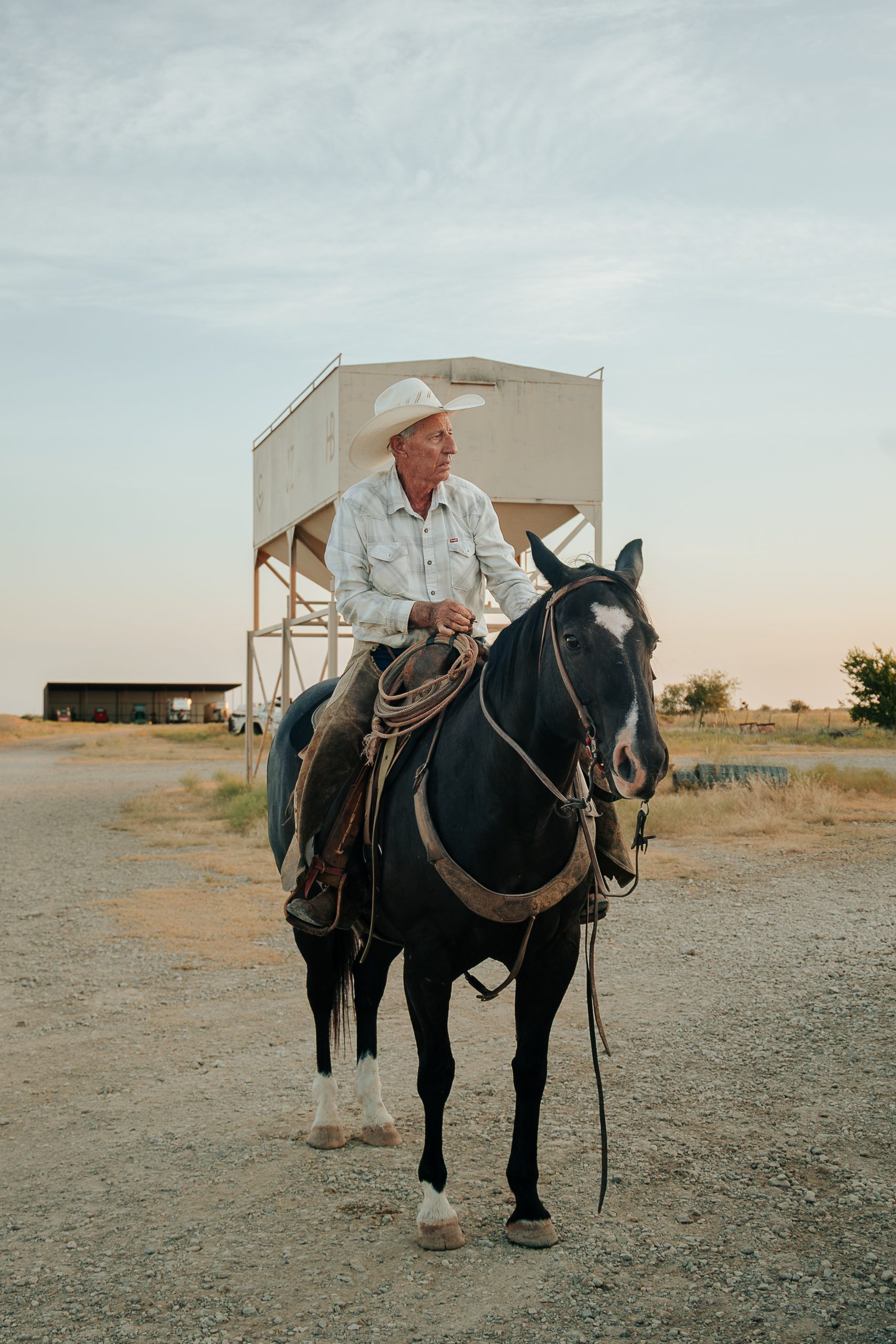 A cowboy in a white hat sits horseback on a black horse with a coiled rope resting on the saddle at a ranch. A feed bin and ranch buildings stand in the background beneath a pale evening sky.