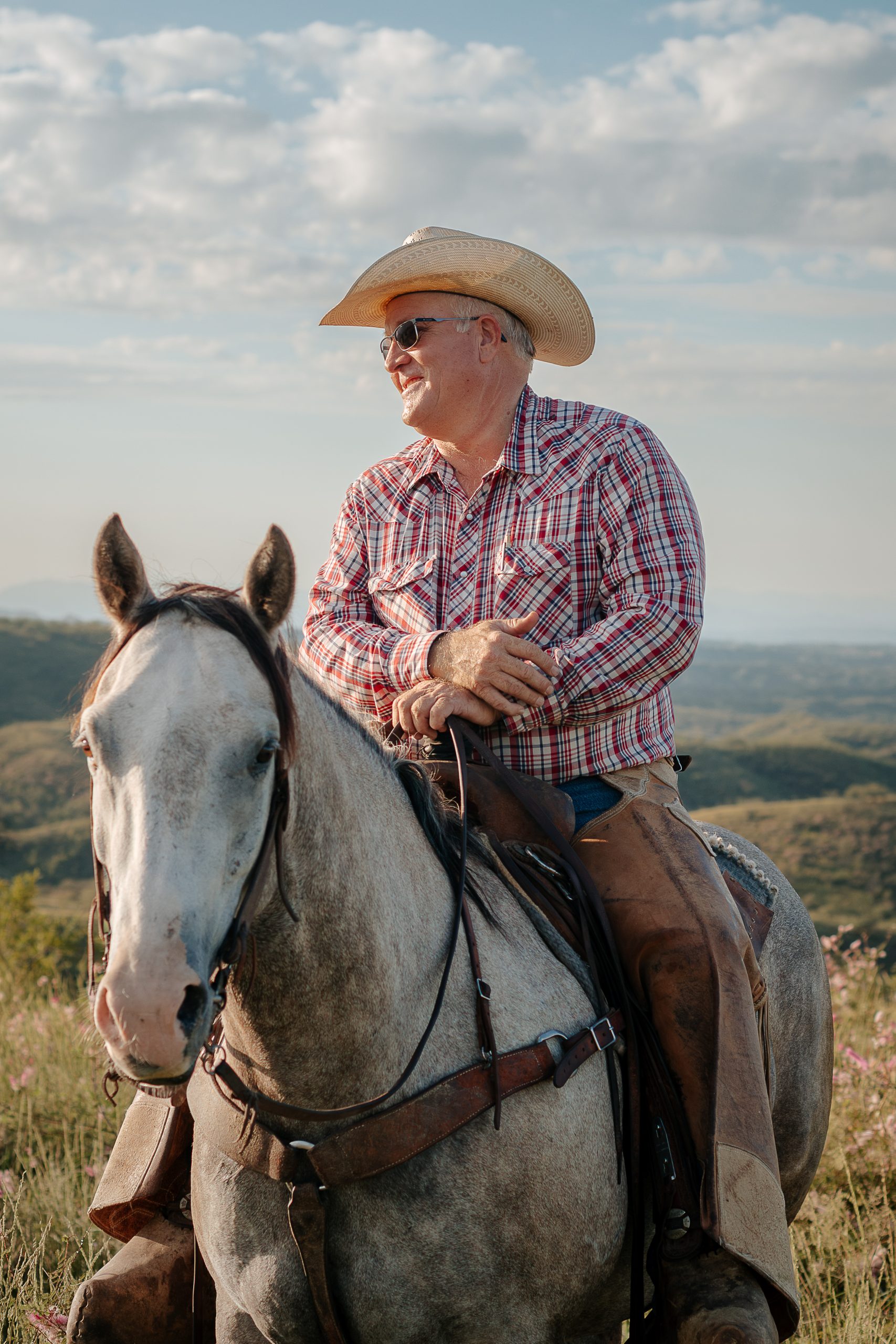Cowboy wearing a straw hat, sunglasses, plaid shirt and leather chaps sits horseback overlooking a rugged ranch landscape.