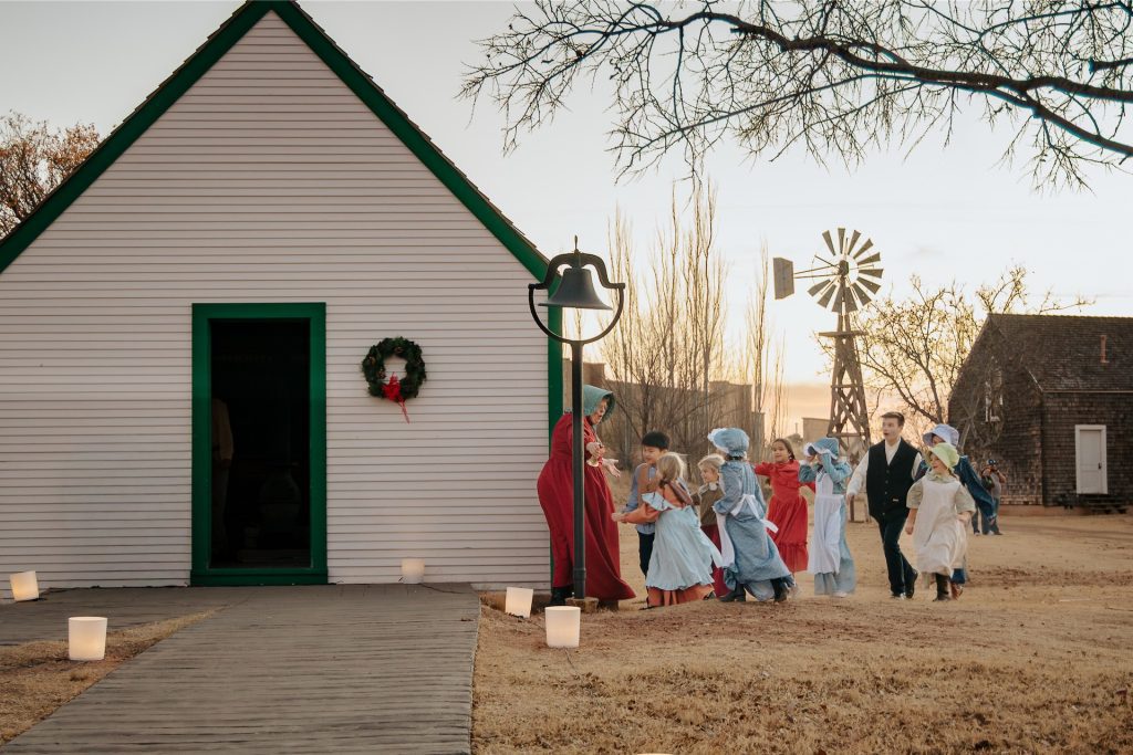 Children and teacher walking toward a one-room schoolhouse.