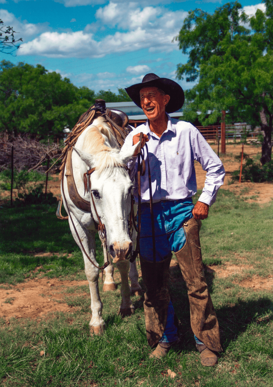 Smiling cowboy in a black hat, blue shirt and leather chaps stands beside a saddled white horse outdoors on a ranch.
