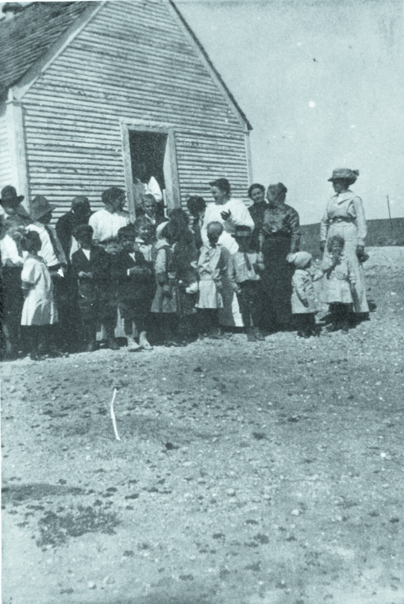 Historic image of children and a teacher in front of the one-room schoolhouse.