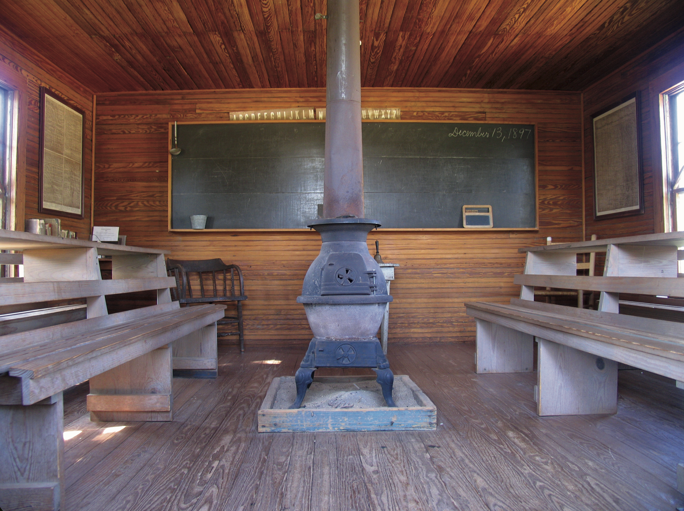 Interior of the Bairfiled Schoolhouse featuring a pot belly stove in the middle, seats on each side and a chalk board at the back.