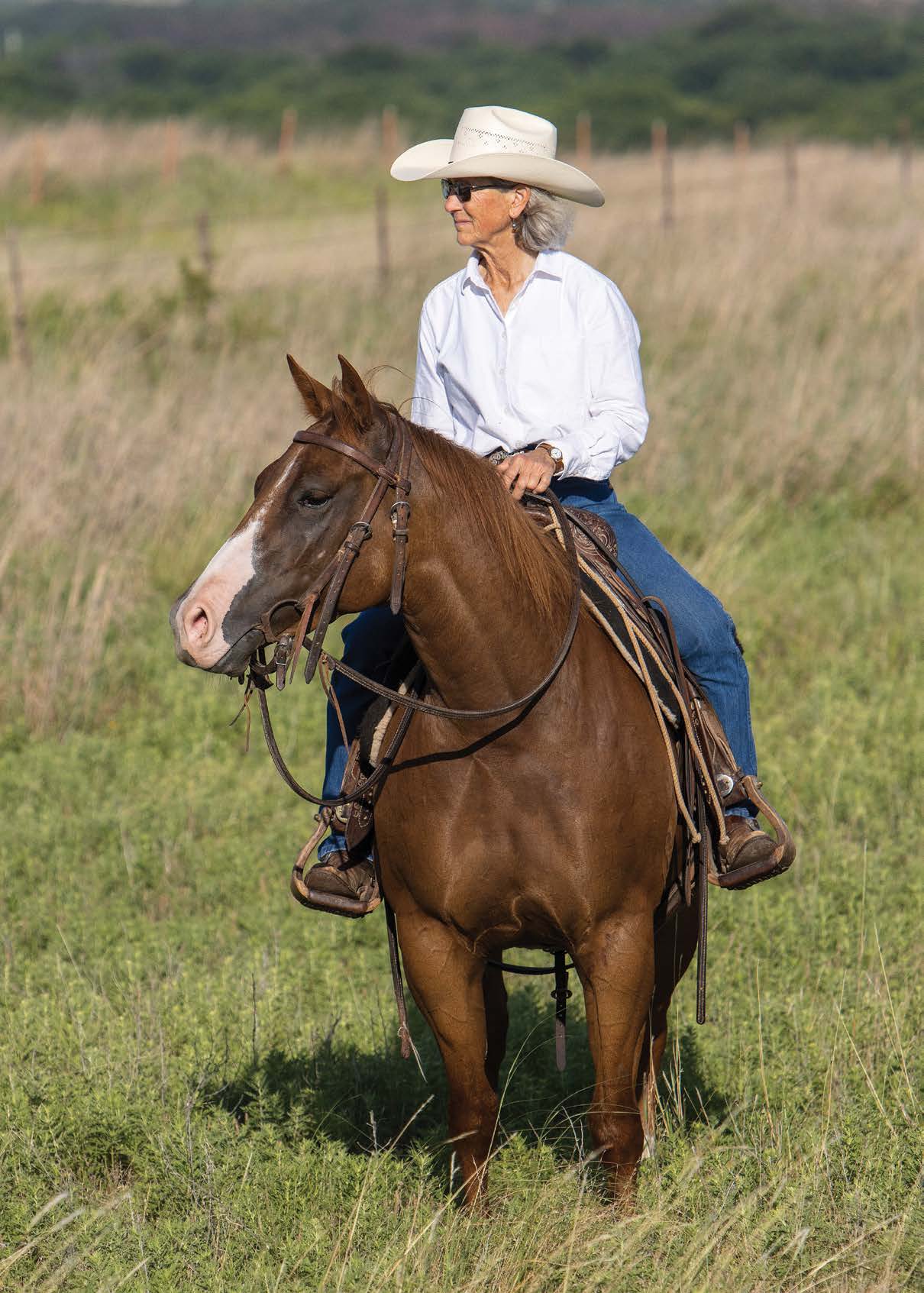 A woman in a white cowboy hat and sunglasses sits horseback in an open pasture surrounded by tall grass.