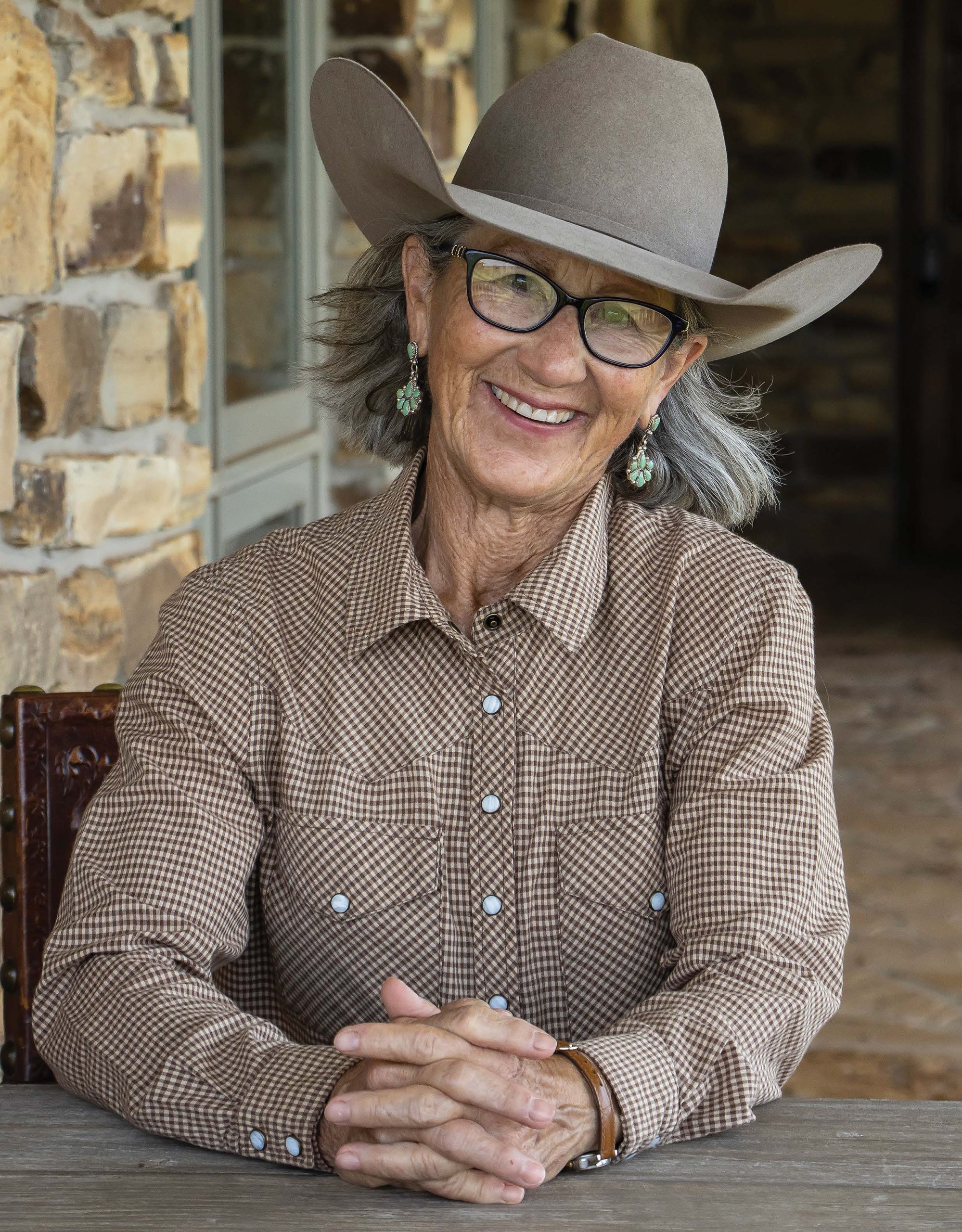 A smiling woman wearing glasses, turquoise earrings and a gray cowboy hat sits at a table in front of a stone building.