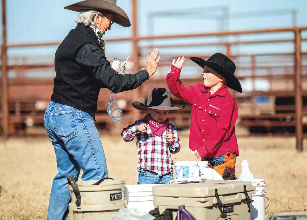 A woman in a cowboy hat high-fives a young child dressed in Western attire while another child stands nearby at an outdoor ranch setting. Coolers and supplies sit in the foreground.