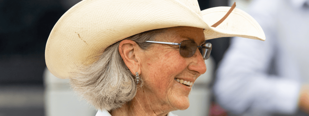 Close-up profile of a smiling woman wearing a white cowboy hat, sunglasses and turquoise earrings.