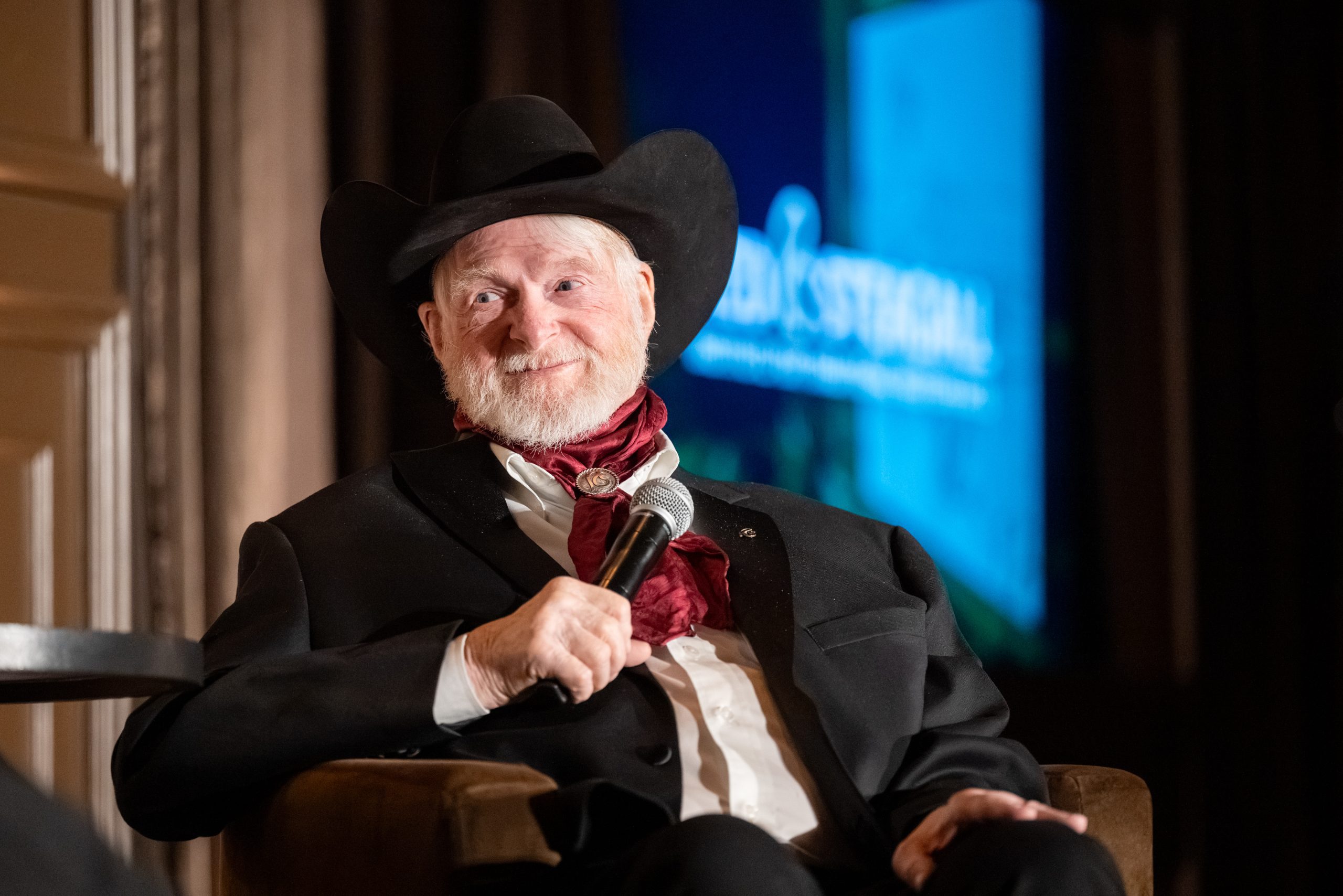 Image of Red Steagall sitting on stage holding a microphone.